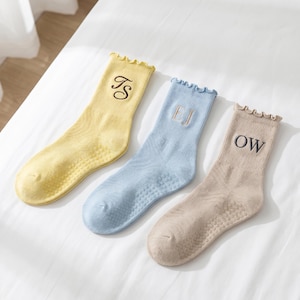 Puede incluir: Tres pares de calcetines personalizados en colores pastel: amarillo, azul claro y beige. Cada calcet&iacute;n presenta una inicial bordada diferente en un color contrastante. Los calcetines tienen un borde superior con volantes y una suela texturizada.