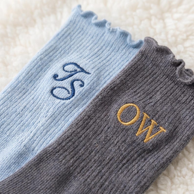 Puede incluir: Dos pares de calcetines acanalados con volantes. Un par es azul claro con las iniciales bordadas en azul oscuro "JS". El otro par es gris con las iniciales bordadas en dorado "OW".