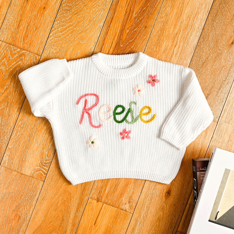 May include: A white knit sweater with the name "Reese" embroidered in pink, green, and yellow. The sweater is decorated with small, embroidered flowers in pink and white. The sweater is lying on a wooden surface.