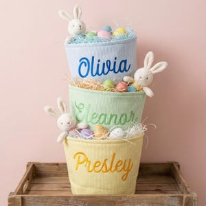 May include: Three pastel-colored Easter baskets stacked on a wooden tray. Each basket has a name embroidered on it: Olivia, Eleanor, and Presley. The baskets are filled with decorative eggs, straw, and a plush bunny.