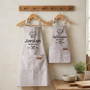 Könnte beinhalten: Zwei gestreifte Schürzen hängen an Holzbügeln. Die Schürzen haben eine Kochmützen-Grafik und die Texte "Jordan Head Chef Est. 1988" und "William Sous Chef Est. 2020". Jede Schürze hat eine Vordertasche.