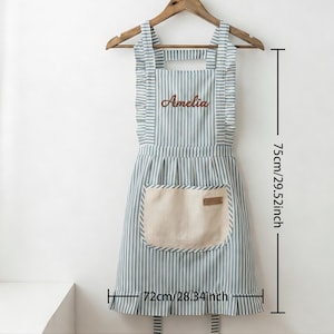 May include: A striped apron in blue and white, with the name "Amelia" embroidered on the chest. It has a beige pocket and measures 29.52 inches in length and 28.34 inches in width. The apron hangs on a wooden hanger.