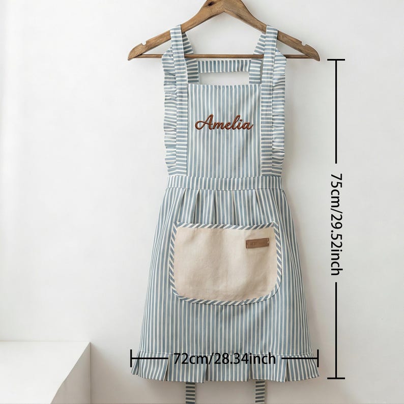 May include: A striped apron in blue and white, with the name "Amelia" embroidered on the chest. It has a beige pocket and measures 29.52 inches in length and 28.34 inches in width. The apron hangs on a wooden hanger.