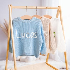 May include: Three knitted sweaters in light blue, beige, and white hang on a wooden rack. The light blue sweater has the name "Lucas" embroidered in white. The beige and white sweaters have embroidered names in white and green.