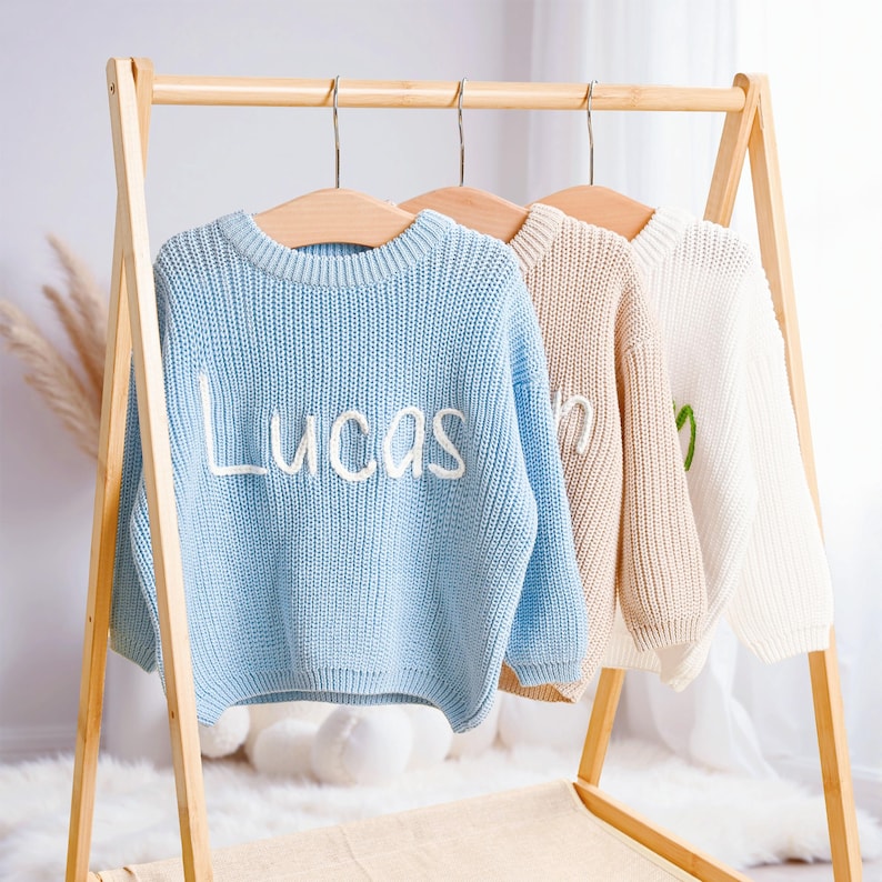 May include: Three knitted sweaters in light blue, beige, and white hang on a wooden rack. The light blue sweater has the name "Lucas" embroidered in white. The beige and white sweaters have embroidered names in white and green.