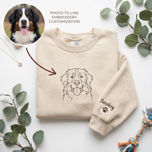May include: A beige crewneck sweatshirt featuring a brown embroidered line art portrait of a Bernese Mountain Dog on the chest. The sleeve is embroidered with the name Smokey and a small paw print. Text above reads PHOTO TO LINE EMBROIDERY CUSTOMIZATION.