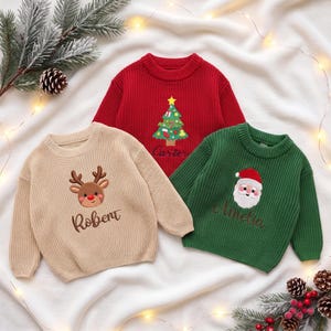 May include: Three long-sleeved knit sweaters in red, beige, and green. The red sweater features a Christmas tree and the name Carter. The beige sweater shows a reindeer and the name Robert. The green sweater displays Santa and the name Amelia.