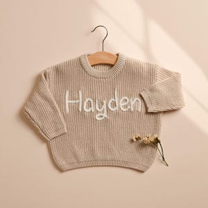 May include: A beige knit sweater with the name "Hayden" embroidered in white script. The sweater is hanging on a wooden hanger. A sprig of dried flowers rests below the sweater.