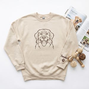 May include: A beige crewneck sweatshirt featuring a dark brown line art illustration of a dog face on the center chest. The left sleeve includes the text "Smokey" in dark brown script lettering above a small dark brown paw print icon.