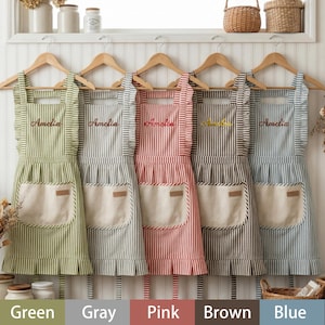 May include: Five striped aprons in green, gray, pink, brown, and blue hang on wooden hangers. Each apron has a large front pocket, ruffled edges, and the name "Amelia" embroidered on the chest. The aprons are made of a striped fabric.
