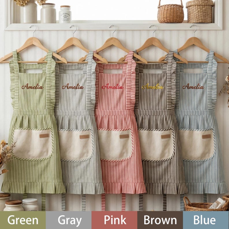 May include: Five striped aprons in green, gray, pink, brown, and blue hang on wooden hangers. Each apron has a large front pocket, ruffled edges, and the name "Amelia" embroidered on the chest. The aprons are made of a striped fabric.