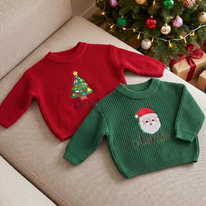May include: A red ribbed knit sweater featuring an embroidered Christmas tree and the name Carter, alongside a green ribbed knit sweater featuring an embroidered Santa Claus face and the name Amelia, both with long sleeves and crew necklines.
