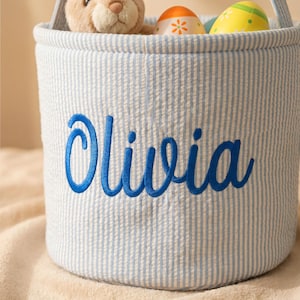 May include: A blue and white striped Easter basket with the name "Olivia" embroidered in blue. The basket contains a stuffed bunny and decorated Easter eggs.