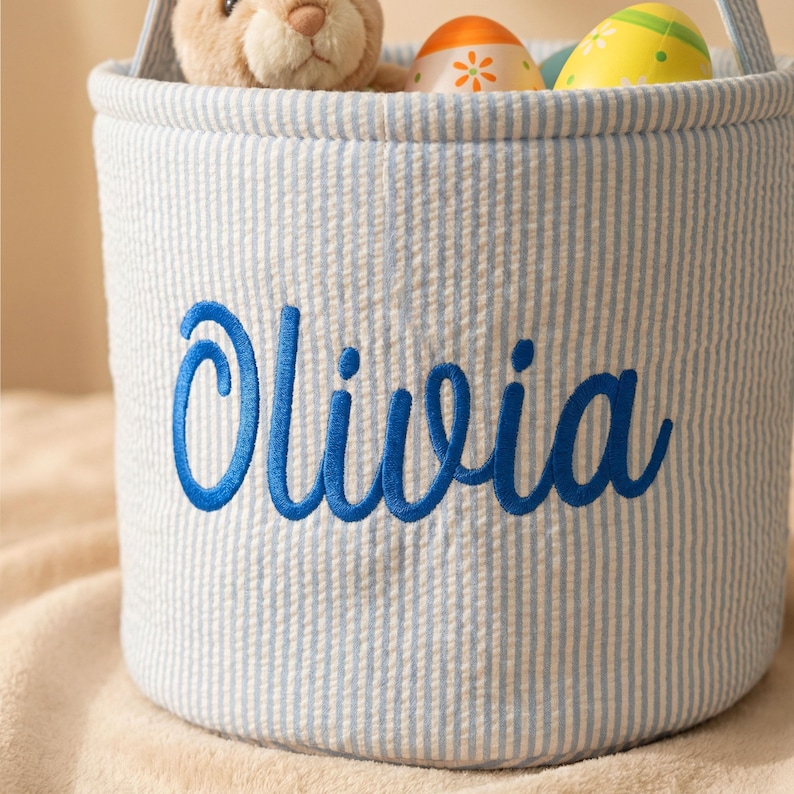 May include: A blue and white striped Easter basket with the name "Olivia" embroidered in blue. The basket contains a stuffed bunny and decorated Easter eggs.