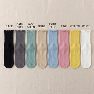 Puede incluir: Una fila de ocho pares de calcetines de colores variados, incluyendo negro, gris oscuro, verde salvia, beige, azul claro, rosa, amarillo y blanco. Cada calcet&iacute;n tiene un borde superior con volantes. Los colores est&aacute;n etiquetados encima de cada calcet&iacute;n.