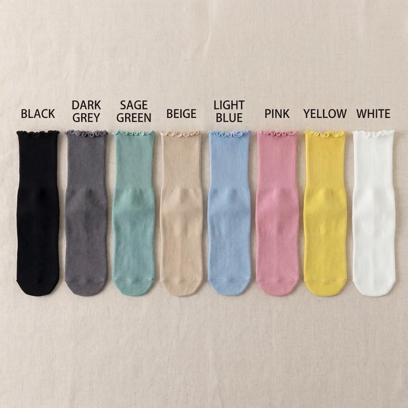 Puede incluir: Una fila de ocho pares de calcetines de colores variados, incluyendo negro, gris oscuro, verde salvia, beige, azul claro, rosa, amarillo y blanco. Cada calcet&iacute;n tiene un borde superior con volantes. Los colores est&aacute;n etiquetados encima de cada calcet&iacute;n.
