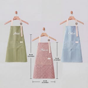 May include: Three striped aprons in green, pink, and blue, each with a name embroidered on the front. The aprons are displayed on wooden hangers. The green apron is 90 cm (35.43 inches) long, the pink apron is 70 cm (27.56 inches) long, and the blue apron is 70 cm (27.56 inches) long.