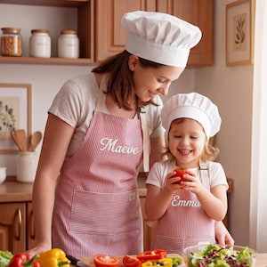 May include: A woman and a child wearing matching red and white striped aprons and white chef hats. The aprons are personalized with the names "Maeve" and "Emma". The child is holding a red tomato. Kitchen setting with vegetables.