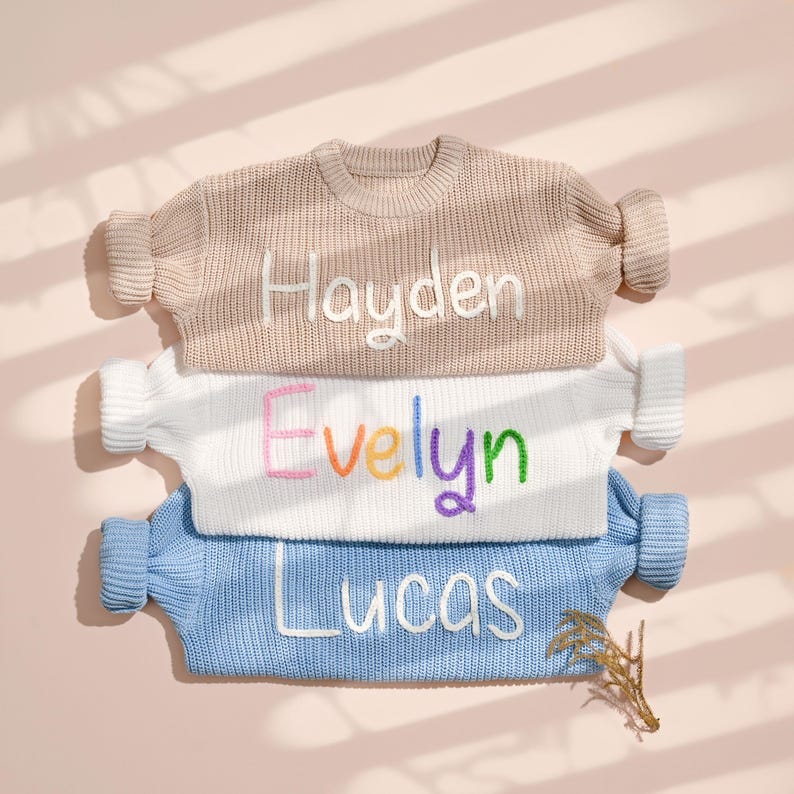 May include: Three stacked children's sweaters in beige, white, and blue. Each sweater has a name embroidered in a contrasting color: "Hayden" in white, "Evelyn" in rainbow colors, and "Lucas" in white. The sweaters are on a pink background.
