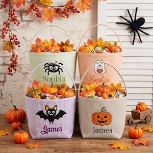 May include: Four striped fabric buckets with handles, each personalized with names Sophia or James and embroidered Halloween icons including a spider, ghost, bat, and jack-o'-lantern. The buckets are filled with artificial autumn leaves and pumpkins.