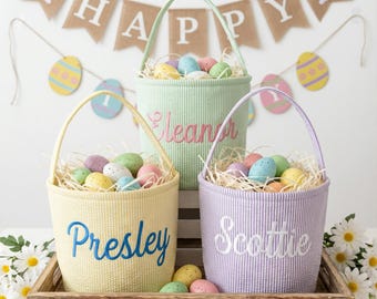 Può includere: Tre cesti pasquali dai colori pastello pieni di uova decorative e paglia, disposti su un vassoio di legno. Ogni cesto ha un manico e un nome ricamato sul davanti: "Eleanor", "Presley" e "Scottie". Uno striscione con "HAPPY" è sullo sfondo.