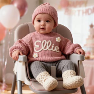 May include: A baby wearing a pink knit sweater with the name "Ellie" in white script, paired with a matching pink beanie, gray leggings, and cream-colored knit booties. The baby is seated in a high chair.
