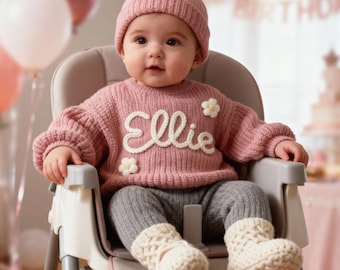 May include: A baby wearing a pink knit sweater with the name "Ellie" in white script, paired with a matching pink beanie, gray leggings, and cream-colored knit booties. The baby is seated in a high chair.
