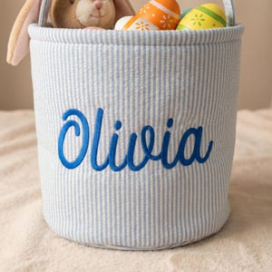 May include: A blue and white striped Easter basket with the name "Olivia" embroidered in blue. The basket contains a stuffed bunny and decorated Easter eggs. The basket has a handle.