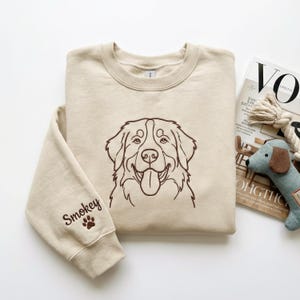 May include: A beige crewneck sweatshirt featuring a brown embroidered line art illustration of a dog face on the center chest. The left sleeve includes the embroidered name Smokey in brown script text accompanied by a small brown paw print icon.
