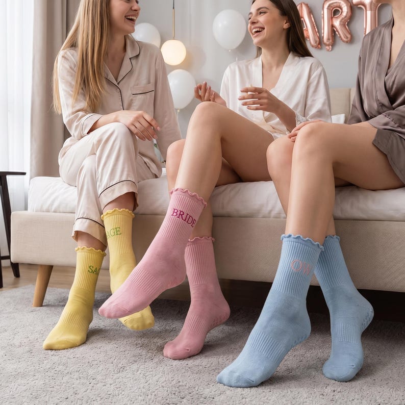 Puede incluir: Tres pares de calcetines deportivos en amarillo, rosa y azul. Los calcetines amarillos tienen la palabra "SAGE" bordada, los rosas "BRIDE" y los azules "OT". Cada par tiene un borde con volantes.