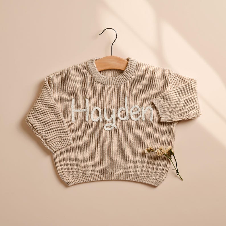 May include: A beige knit sweater with the name "Hayden" embroidered in white script. The sweater is hanging on a wooden hanger. A sprig of dried flowers rests below the sweater.