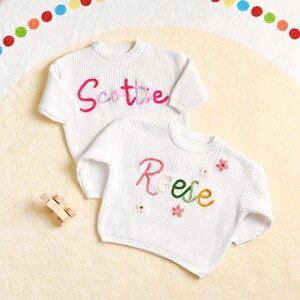 May include: Two white knit sweaters with embroidered names and floral accents. One sweater says "Scottie" in pink and purple thread. The other says "Reese" in pink, green, and yellow thread, with small flower details. A small wooden toy car is in the corner.