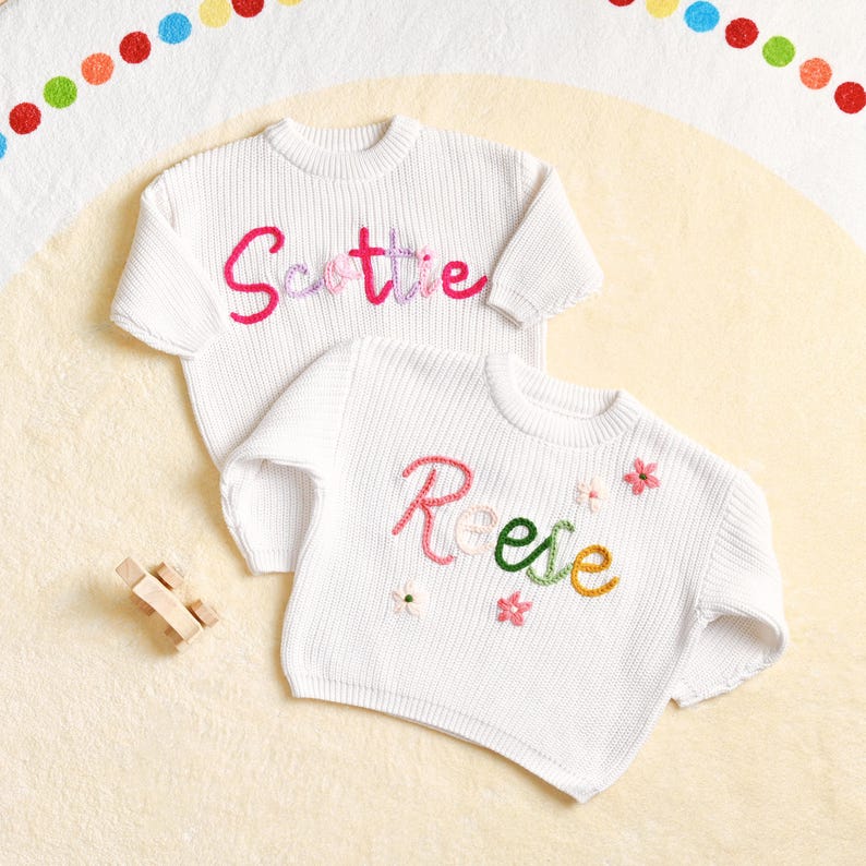 May include: Two white knit sweaters with embroidered names and floral accents. One sweater says "Scottie" in pink and purple thread. The other says "Reese" in pink, green, and yellow thread, with small flower details. A small wooden toy car is in the corner.