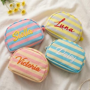 Puede incluir: Cuatro neceseres a rayas en colores pastel, cada uno bordado con un nombre diferente: Stella, Luna, Victoria y Daisy. Los neceseres tienen forma redondeada y un pequeño tirador de cremallera con pompón.