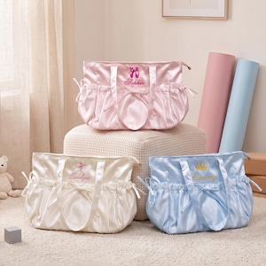 May include: Three satin tote bags in light pink, cream, and light blue. Each bag features two handles, side drawstring ties, and custom embroidery. The pink bag displays ballet slippers and the name Sophie, the cream bag a dancer and Evelyn, and the blue bag a crown and Rosemary.