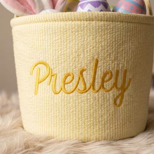 May include: A yellow Easter basket with the name "Presley" embroidered in gold script. The basket is filled with colorful Easter eggs and a pair of pink bunny ears.