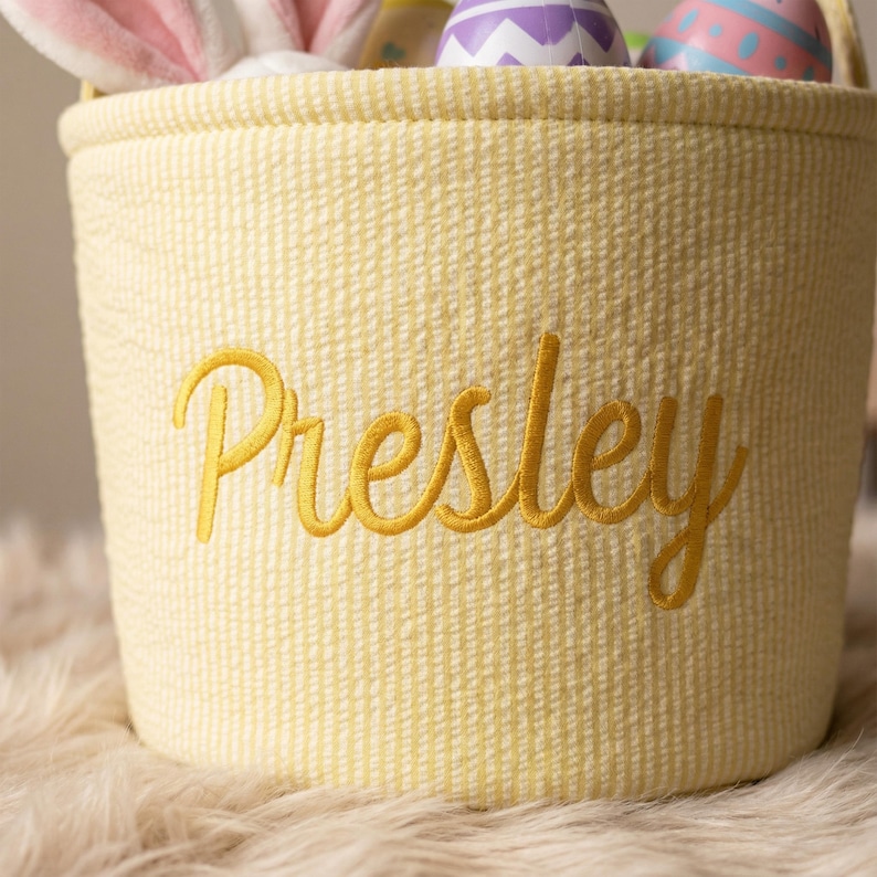 May include: A yellow Easter basket with the name "Presley" embroidered in gold script. The basket is filled with colorful Easter eggs and a pair of pink bunny ears.
