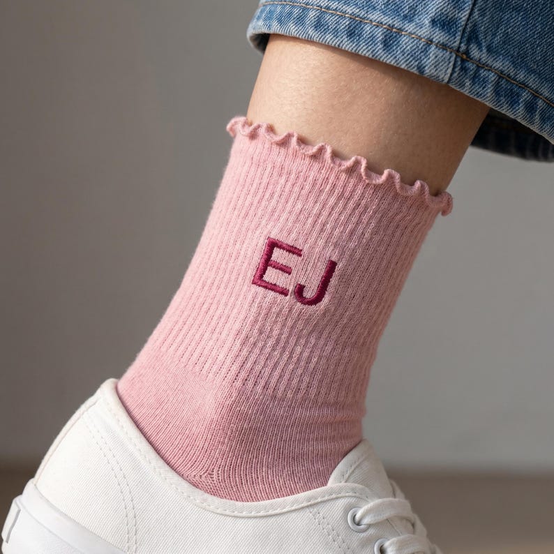 Puede incluir: Calcetines rosas de canal&eacute; con un borde festoneado y las letras "EJ" bordadas en rosa oscuro. Los calcetines se llevan con zapatillas blancas y vaqueros azules.