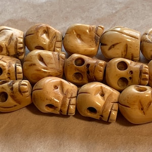 May include: Close-up of a collection of carved skull beads. The beads are a light tan color with detailed facial features and carved lines. The beads have a smooth, polished finish and are arranged closely together.