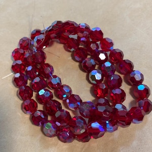 May include: Strand of faceted, deep red beads with iridescent highlights. The beads are strung together, forming a circular shape. The beads are a deep red color and have a faceted cut that catches the light.