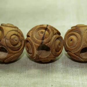 May include: Three intricately carved wooden spheres. Each sphere is a warm brown color and features a series of concentric circles and geometric patterns. The spheres are evenly spaced, showcasing the detailed craftsmanship.