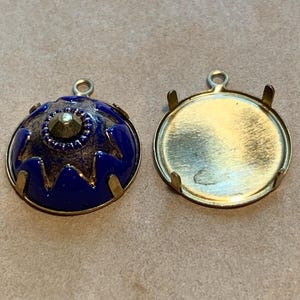 May include: Two ornate pendant settings. One is a deep blue, domed shape with a central gem and gold accents. The other is a gold-toned, open-backed setting with prongs for securing a stone or embellishment. Both have small loops for hanging.