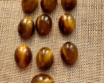 Könnte beinhalten: Eine Sammlung von elf polierten Tigeraugen-Edelsteinen. Die oval geformten Steine haben eine glatte, reflektierende Oberfläche. Sie sind warmes Goldbraun mit dunkleren braunen Streifen, die einen gestreiften Effekt erzeugen. Die Edelsteine sind auf einer strukturierten, beigefarbenen Oberfläche angeordnet.
