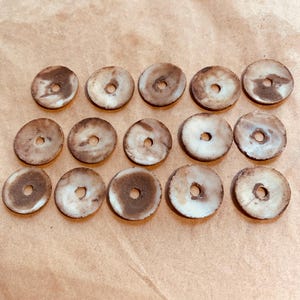 Peut inclure: Une collection de quinze perles rondes et plates avec un trou central. Les perles présentent un aspect marbré avec des nuances de marron et de crème. Elles sont disposées en grille sur une surface neutre.
