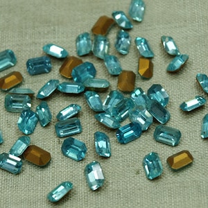 May include: A large quantity of turquoise blue rectangular glass rhinestones. The rhinestones are scattered on a light blue fabric surface.