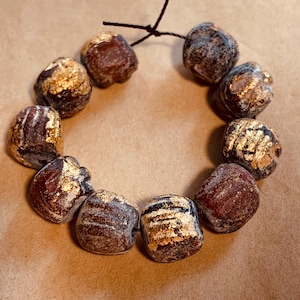 May include: A handmade bracelet featuring a series of irregularly shaped beads. The beads are primarily dark red and brown, with gold leaf accents. The bracelet is strung on a black cord, creating a unique and artistic accessory.