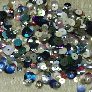 May include: A large assortment of iridescent sequins in various colors, including blue, green, white, and pink. The sequins are scattered on a light brown fabric surface.