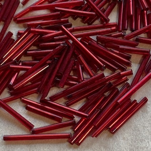May include: A collection of small, cylindrical, red glass beads scattered across a surface. The beads are a deep, translucent red, reflecting light and creating highlights. These beads are suitable for jewelry making and craft projects.