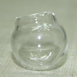 May include: A clear glass jar with a round shape and a slightly uneven opening. The jar is sitting on a textured surface.