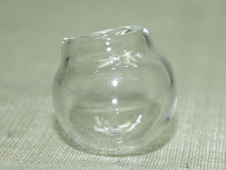 May include: A clear glass jar with a round shape and a slightly uneven opening. The jar is sitting on a textured surface.